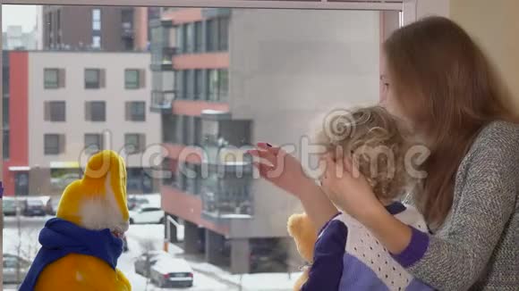 妈妈带着蹒跚学步的小女孩坐在靠近窗户的地方享受雪花般的雪花飘落视频的预览图