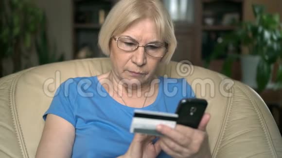 专心致志的老年妇女坐在家里的椅子上用信用卡在线购买智能手机视频的预览图