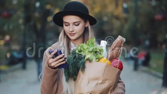 年轻漂亮的女人穿着时髦的外套站在秋天的公园里拿着一包产品用智能手机视频的预览图