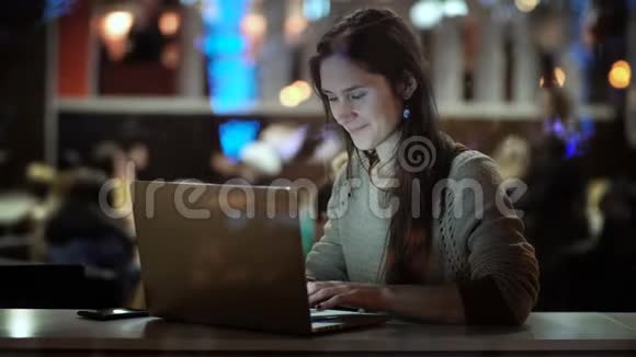 在窗口内查看年轻快乐的女人晚上坐在咖啡馆里用笔记本电脑微笑着视频的预览图