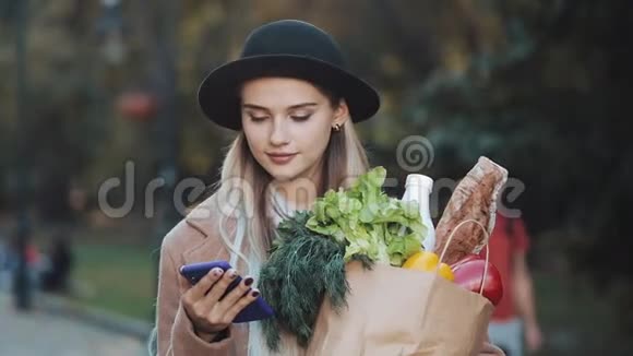 年轻漂亮的女人穿着时髦的外套站在秋天的公园里拿着一包产品用智能手机视频的预览图