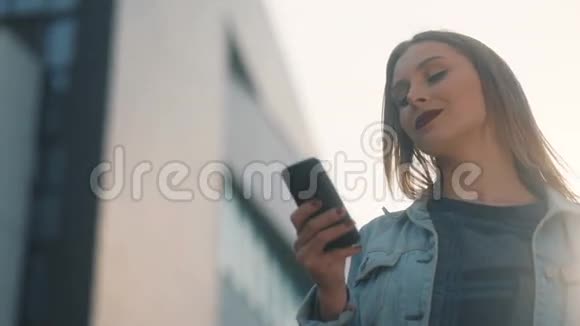 在蓝牛仔布球场用她的手机拍摄的带有暗红色嘴唇的金发女孩的都市化镜头以及严格的模糊镜头视频的预览图
