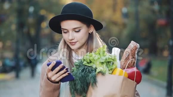 年轻漂亮的女人穿着时髦的外套站在秋天的公园里拿着一包产品用智能手机视频的预览图