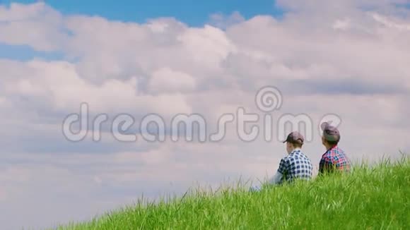两个戴着帽子的十几岁的男孩坐在一座绿色的小山上靠着蓝天他们说话向天空举手儿童视频的预览图