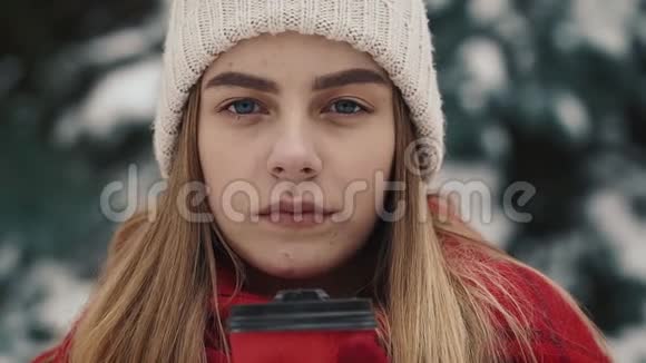 年轻漂亮的女孩穿着温暖的衣服站在圣诞树附近在雪地里喝着报纸上的热饮料视频的预览图