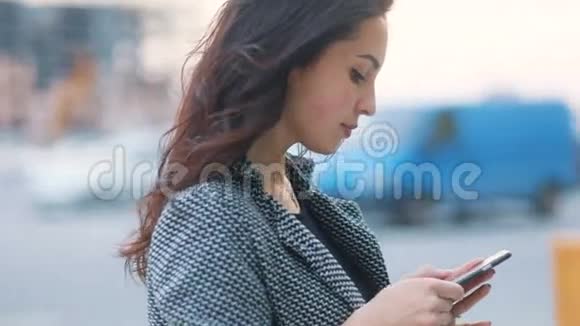 穿着严格的灰色夹克和黑色连衣裙的年轻温柔的商务女孩在繁忙的城市中停留的侧视特写视频的预览图
