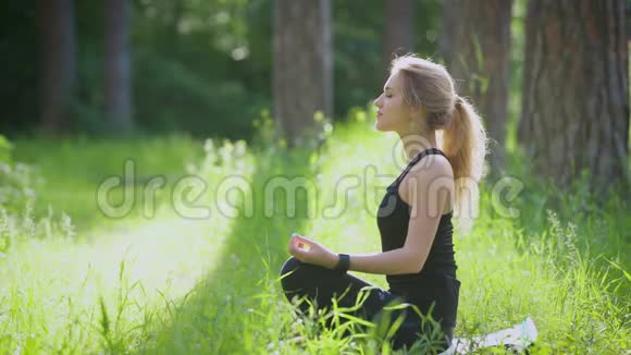 年轻的金发女人穿着运动服在大自然中冥想女人坐在莲花的位置双手跪在绿草上视频的预览图