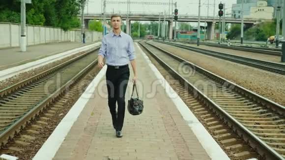 一位年轻成功的商人沿着铁轨沿着火车站散步他手里拿着一个包微笑着A视频的预览图