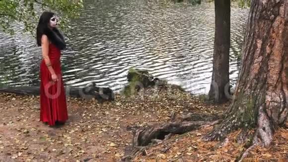 一个穿着红色衣服的女人穿着恐怖的万圣节服装在森林公园的树林间散步视频的预览图