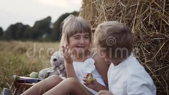 一个穿着白色裙子的小女孩吵醒了一个正在吃李子的男孩田野里的孩子们坐在干草捆下视频的预览图