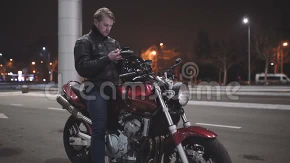 一个穿皮夹克的年轻人坐在摩托车上在夜街打电话视频的预览图