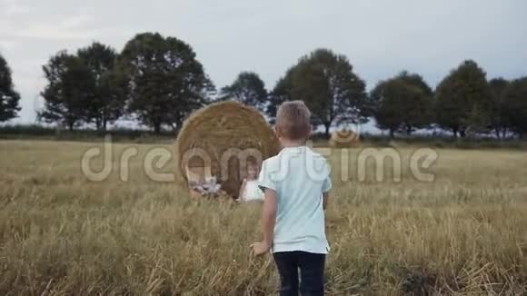 一个小男孩跑到一个女孩面前她坐在稻草堆下的田野里快乐的女孩穿着白色的衣服微笑的男孩奔跑视频的预览图