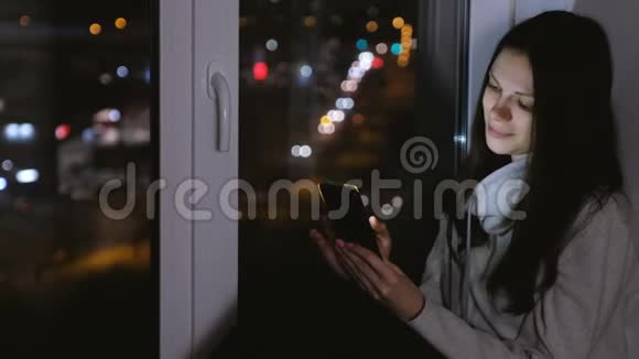 女人在黑暗的夜晚坐在窗台上用手机浏览网页道路背景视频的预览图
