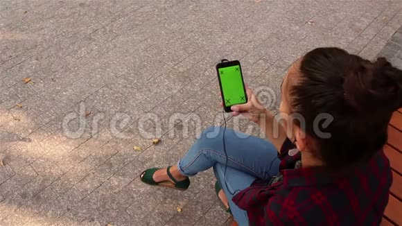 年轻漂亮的女孩在智能手机上听音乐坐在公园的长凳上视频的预览图