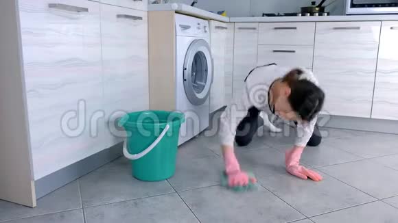穿着粉红色橡胶手套的疲惫的女人用一块布洗厨房的地板地板上的灰色瓷砖视频的预览图