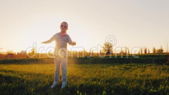 穿着粉红色眼镜和运动服的有趣女孩做体操日落时分阳光穿过绿色的草地视频的预览图