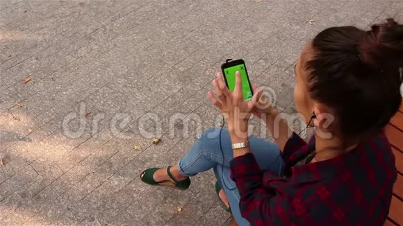 年轻漂亮的女孩在智能手机上听音乐坐在公园的长凳上视频的预览图