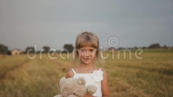美丽的女孩一头长发穿着白色连衣裙带着一只柔软的玩具熊走在田野视频的预览图