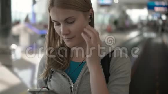 快乐微笑的女人在机场候机楼里用牵引器使用她的智能手机听音乐视频的预览图