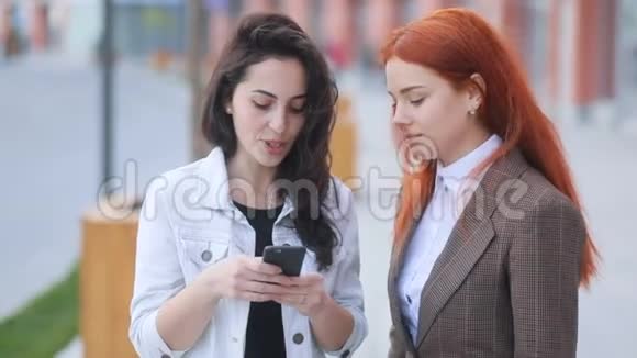 两位年轻迷人的商业女性红发和棕色头发在智能手机中说话和表演动作视频的预览图