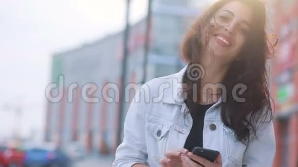 漂亮的棕色头发的白种人女孩呆在购物中心前的阳光下用她的智能手机看着视频的预览图