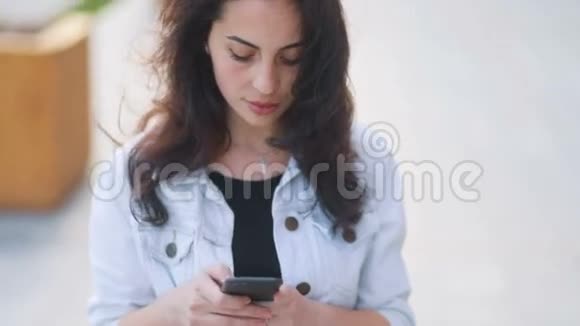 穿着时髦的白色牛仔裤外套的棕色头发的令人惊叹的白种人女孩与智能手机交流随风摇曳的头发视频的预览图