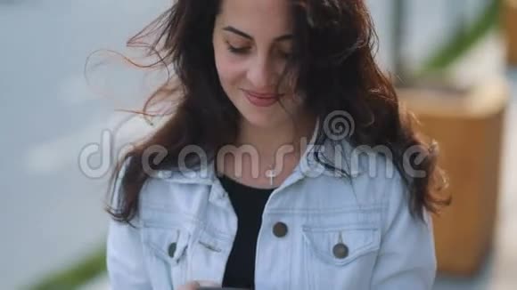 棕色头发的白种人女孩带着迷人的微笑穿着浅白色牛仔裤外套用她的智能手机看着她的背影视频的预览图