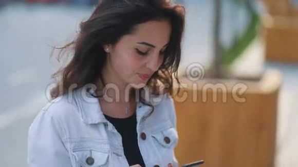 年轻漂亮的白种人棕色头发的女孩穿着浅蓝色牛仔裤外套在都市背景下使用她的智能手机视频的预览图
