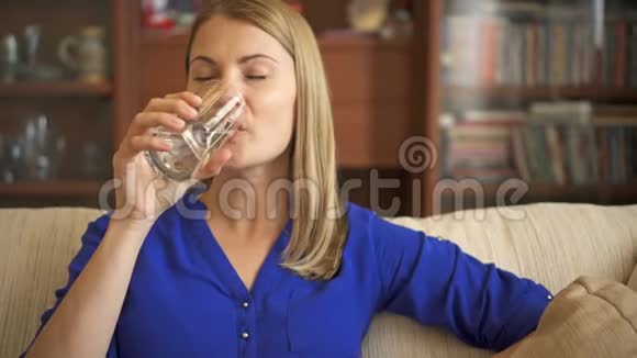 一位美丽迷人的女士穿着蓝色衬衫坐在客厅的沙发上用玻璃喝水视频的预览图