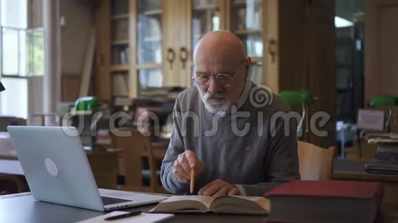 成熟的教授用书和笔记本电脑工作坐在图书馆的桌子旁视频的预览图