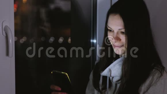 女人在黑暗的夜晚坐在窗台上用手机浏览网页道路背景视频的预览图