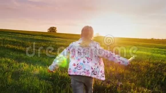 一个无忧无虑的女孩跑过绿色的田野走向日落概念快乐的童年光明和自由视频的预览图