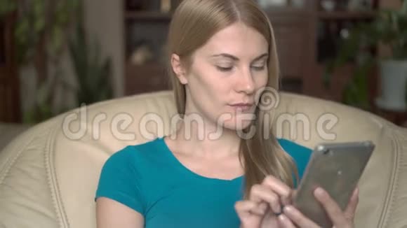 有趣的积极的年轻女人坐在家里的椅子上利用她的智能手机浏览阅读新闻视频的预览图
