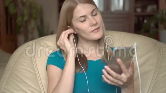 有趣开朗的女人坐在家里的椅子上在智能手机上用耳机听音乐唱歌视频的预览图