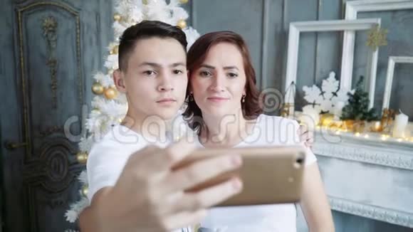 一个年轻人带着他美丽的母亲在圣诞树的背景下在智能手机上自拍视频的预览图
