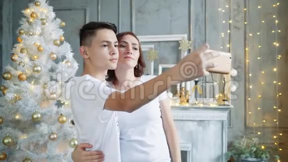 一个年轻人带着他美丽的母亲在圣诞树的背景下在智能手机上自拍视频的预览图