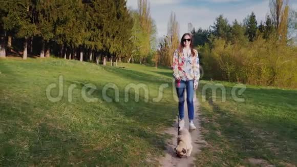 带着太阳镜和狗在公园散步的年轻女人沿着这条小路走在风景如画的大自然的背景下视频的预览图