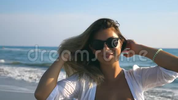 一个穿着比基尼和衬衫的快乐女孩在海岸边散步微笑着戴墨镜的年轻美女视频的预览图