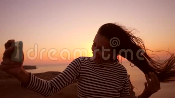 年轻的混合种族女孩使用手机拍摄自拍照片在惊人的日落背景高清慢速运动视频的预览图