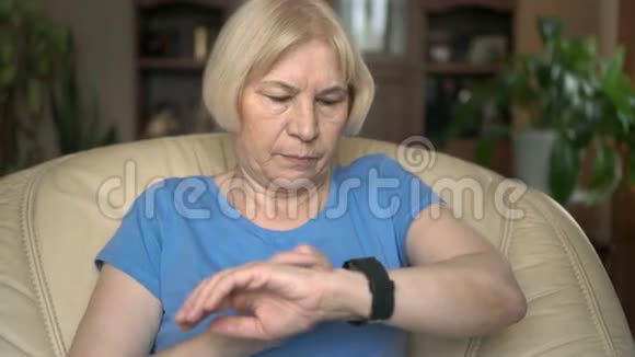 现代老年妇女坐在黄色的椅子上用她的黑色智能手表浏览阅读视频的预览图