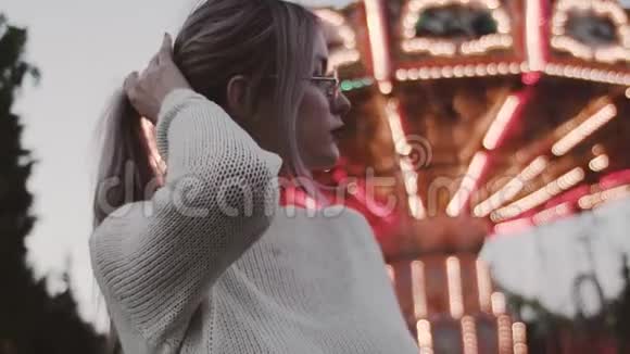 戴眼镜的漂亮女孩在游乐园附近抚摸着旋转木马的头发视频的预览图