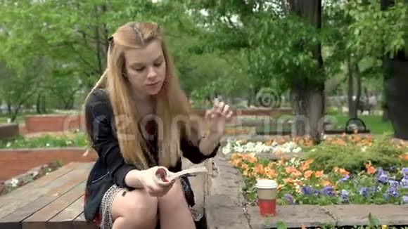 美丽的金发女郎在夏天的公园里喝咖啡读书视频的预览图