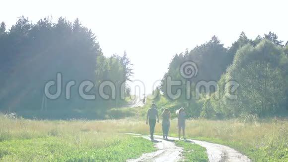 两个女孩和一个男人在夏天的时候在日落的好天气里沿着乡间小路和森林散步视频的预览图