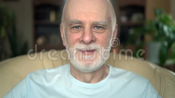 家中坐在椅子上的英俊微笑老人的肖像在镜头前眨眼视频的预览图