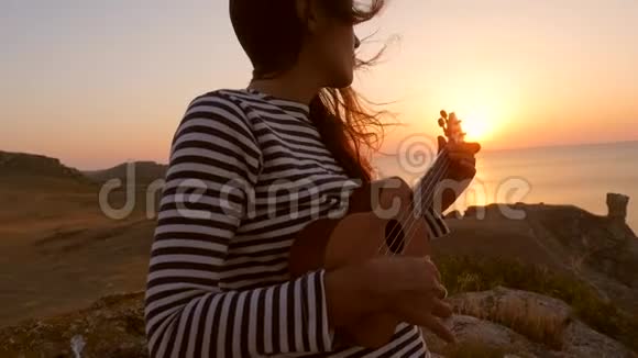 水手嬉皮白种人女孩演奏四弦琴吉他在美丽的日落附近的海洋高清慢镜头视频的预览图
