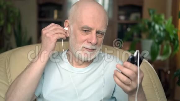 好看的老人坐在家里的椅子上在智能手机上用耳机听音乐唱歌视频的预览图