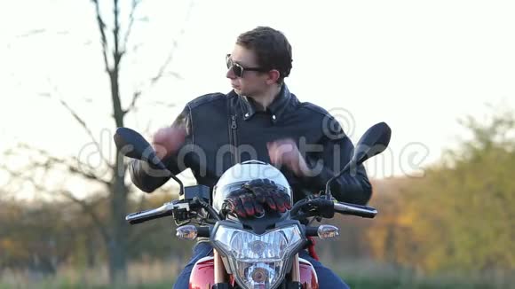 一个穿着黑色皮夹克的年轻人坐在摩托车上戴上太阳镜戴上白色头盔然后在秋日旅行视频的预览图