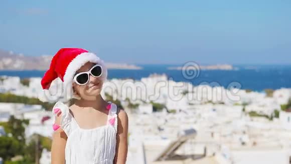戴着圣诞帽的小女孩在户外背景是Mykonos的老街典型的希腊传统村落街上的孩子视频的预览图