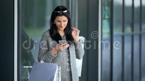 漂亮的年轻时尚女士在商店橱窗附近有购物袋打着智能手机视频的预览图