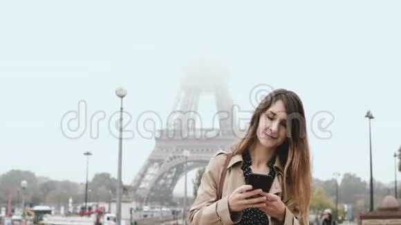 年轻漂亮的女人站在法国巴黎埃菲尔铁塔附近在雾蒙蒙的早晨使用智能手机视频的预览图